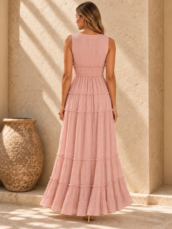 A woman stands with her back to the camera wearing a Villa Vibes sleeveless, tiered maxi dress in dusty pink. The dress features a textured crinkle fabric and delicate ruffle trim at the waist and between the tiers.