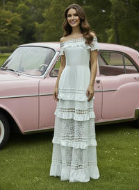 Woman in a white Alexa lace dress standing in front of a pink vintage car on grass.