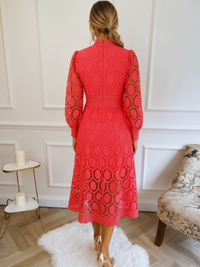 A woman viewed from behind wearing a Villa Vibes Jasmine bright coral-red midi dress with a high mock neck, long sleeves, and an allover circular lace pattern. She is standing indoors on a white rug in front of a white paneled wall, with a small side table and a patterned armchair visible.