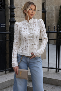 Woman wearing a white lace top and blue jeans, holding a YSL handbag, standing in an urban setting.