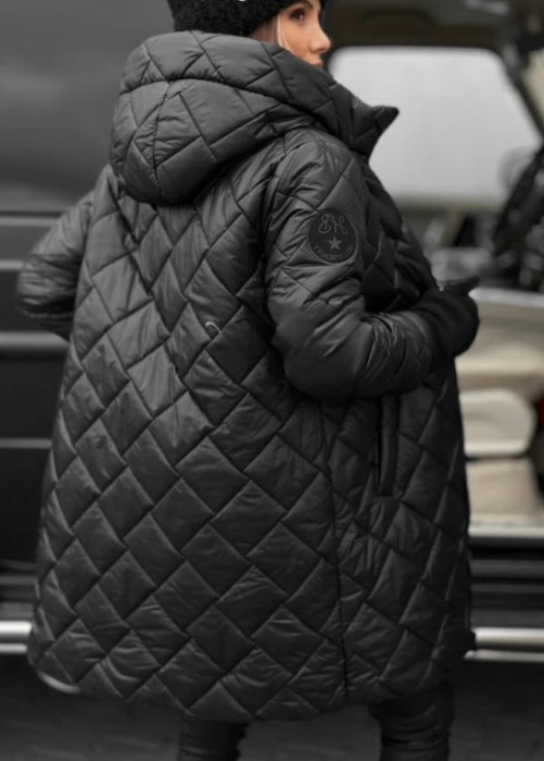 Person wearing a black quilted coat with a brand logo, standing in front of a blurred background.