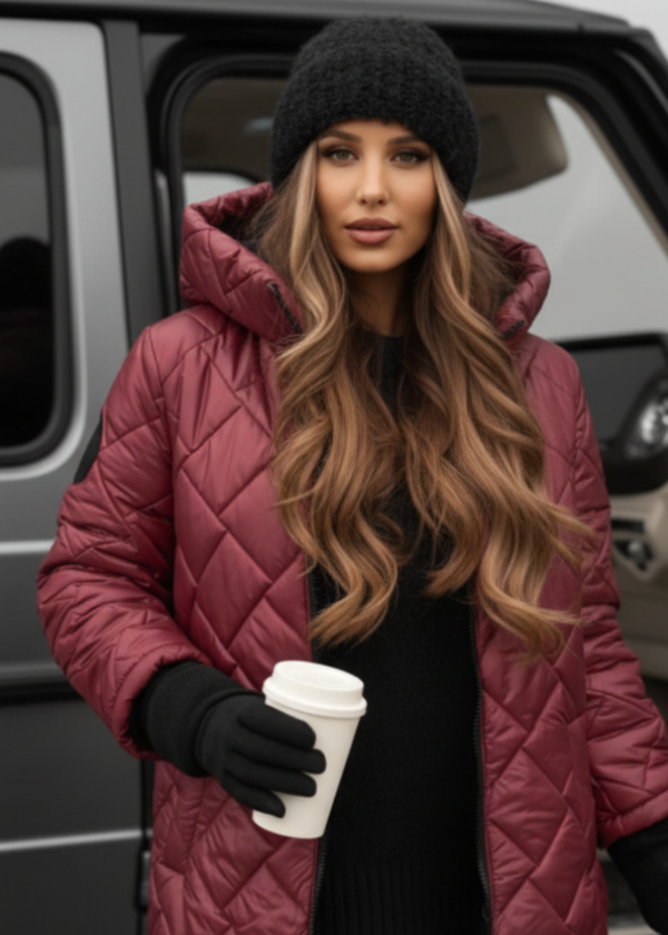 Woman in a maroon quilted jacket and black beanie holding a white cup, standing in front of a vehicle.