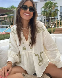 Woman wearing a Villa Vibes white embroidered top sitting outdoors with a pool and palm trees in the background.