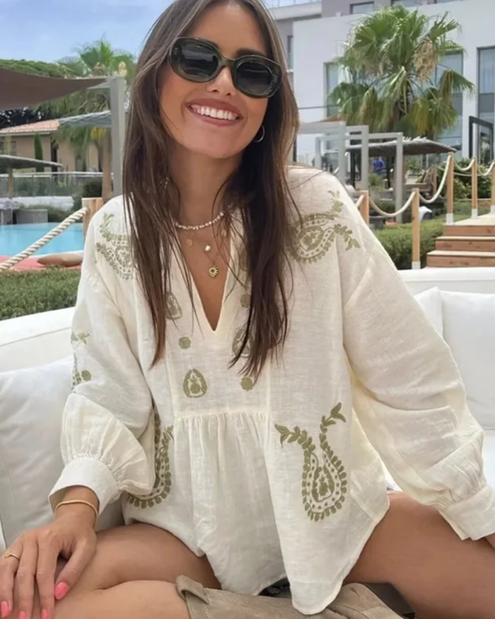 Woman wearing a Villa Vibes white embroidered top sitting outdoors with a pool and palm trees in the background.