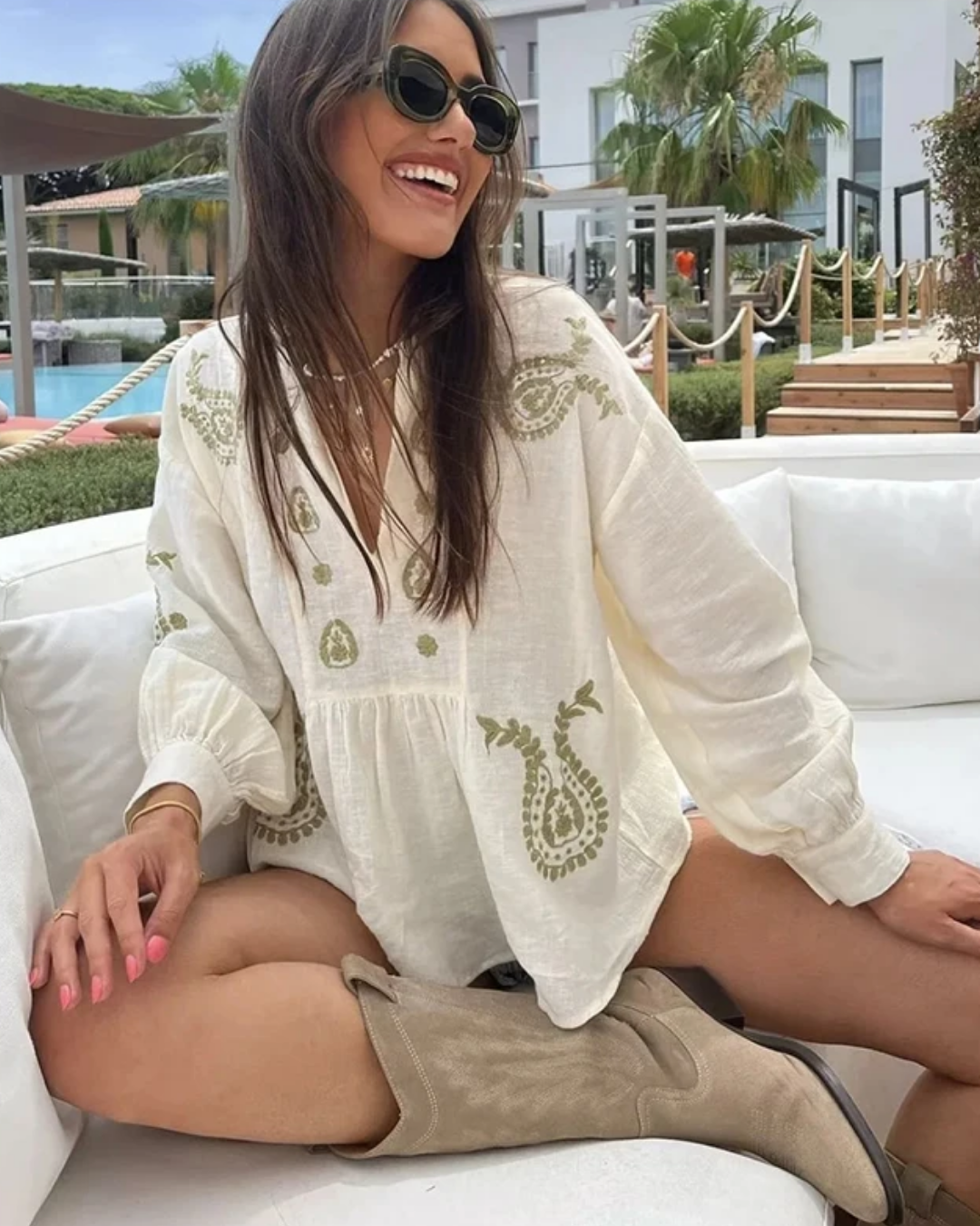 Woman sitting on a white couch wearing a Villa Vibes white embroidered blouse and brown boots, with a pool and palm trees in the background.