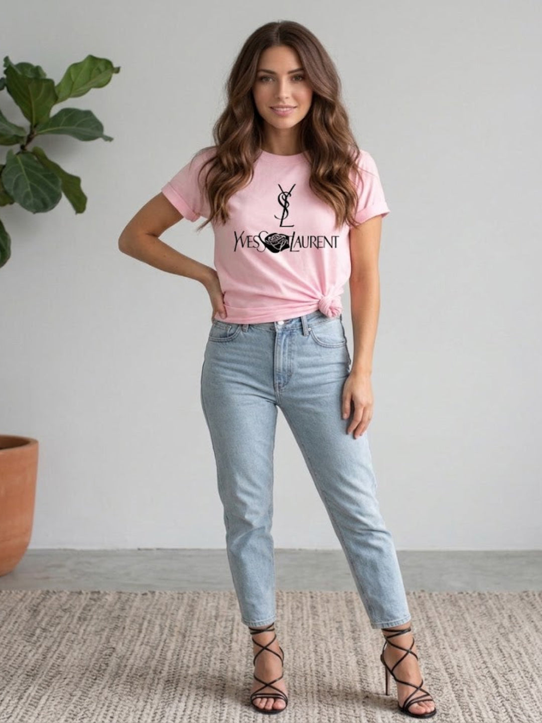 A woman modeling a Villa Vibes Designer Inspired light pink graphic t-shirt with a black Yves Saint Laurent logo, paired with light-wash blue jeans and black lace-up heels, standing in a minimalist room with a potted plant.