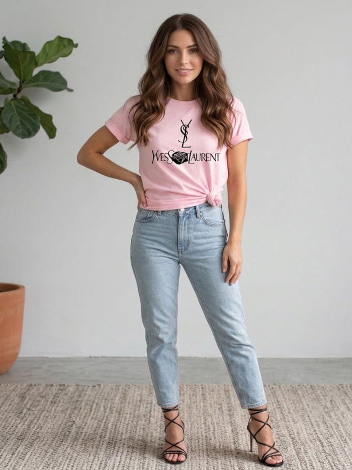 A woman modeling a Villa Vibes Designer Inspired light pink graphic t-shirt with a black Yves Saint Laurent logo, paired with light-wash blue jeans and black lace-up heels, standing in a minimalist room with a potted plant.