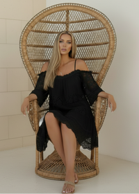 Woman in a Fallon black dress sitting on a wicker peacock chair against a white wall.