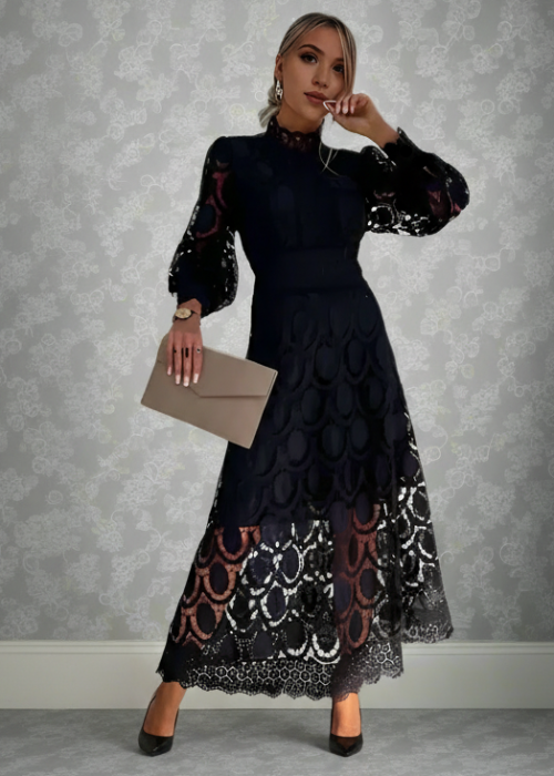 Woman in a black lace dress standing against a gray patterned wall.