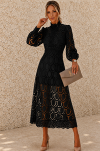 A woman models a black midi dress with long sheer lace sleeves and a circular lace overlay skirt, holding a taupe clutch against a textured beige wall.
