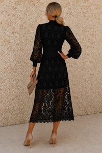 A woman shown from the back wearing a black midi dress with sheer lace sleeves and a scalloped lace hem, standing against a textured beige wall.