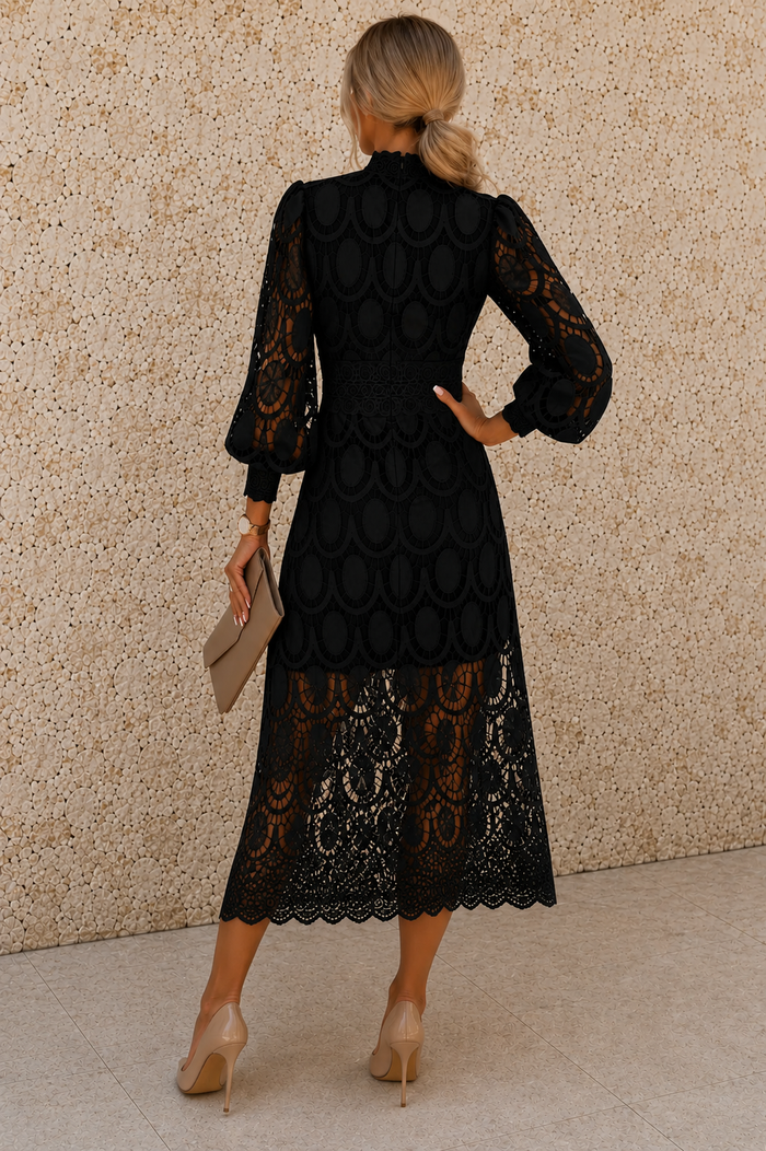 A woman shown from the back wearing a black midi dress with sheer lace sleeves and a scalloped lace hem, standing against a textured beige wall.
