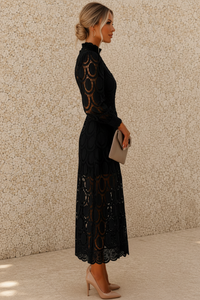 A woman in a black lace long-sleeve midi dress stands in profile against a textured beige wall, holding a taupe clutch and wearing beige heels.