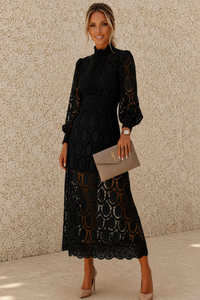 A woman models a black long-sleeve midi dress with a high mock neck and a scalloped lace hem, holding a taupe clutch bag against a textured beige wall.