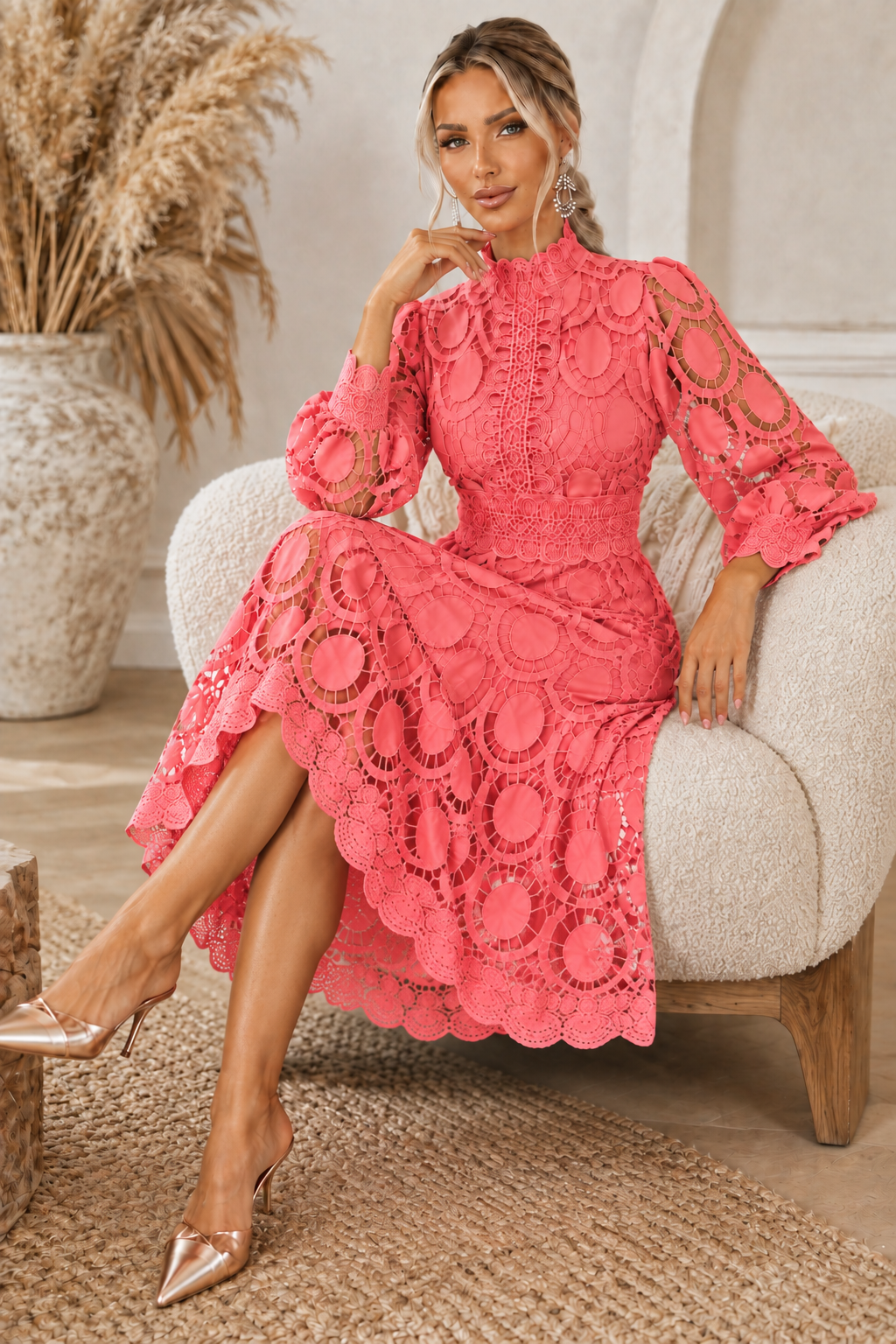 A woman models a Jasmine coral pink lace midi dress with long sleeves, a high mock neck, and circular lace patterns, sitting on a cream-colored armchair in a styled room.