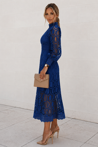 Woman wearing a blue lace dress holding a beige clutch against a white background