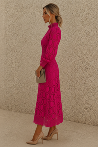 A woman in profile wearing a bright pink long-sleeve midi dress with a circular lace pattern, holding a small taupe clutch and wearing beige heels, standing against a textured beige wall.