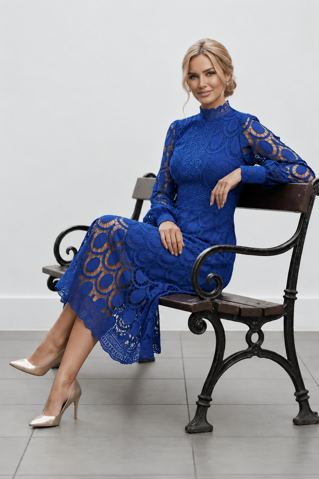 Woman in a blue lace dress sitting on a black metal bench against a white wall.