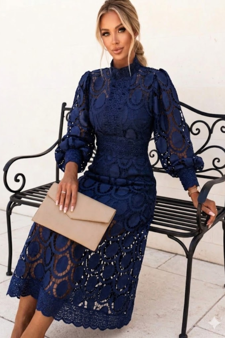 Woman in a blue Jasmine lace dress sitting on a bench holding a beige clutch.