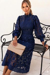 Woman in a blue Jasmine lace dress sitting on a bench holding a beige clutch.