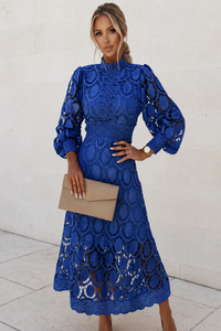 Woman wearing a blue lace dress holding a beige clutch against a white background