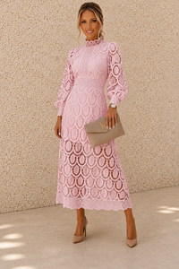 A woman models a light pink long-sleeve lace midi dress with a high ruffled collar and an allover circular lace pattern. She is holding a taupe clutch and wearing nude heels, standing against a textured beige stucco wall.