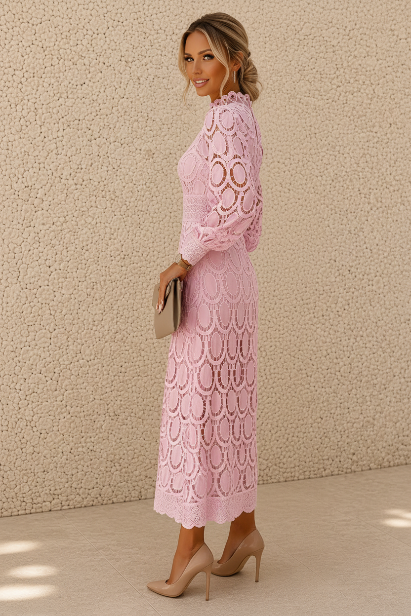A woman modeling a light pink long-sleeve lace midi dress with a ruffled high neckline, standing against a textured beige stucco wall.