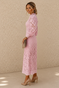 A woman modeling a light pink long-sleeve lace midi dress with a ruffled high neckline, standing against a textured beige stucco wall.