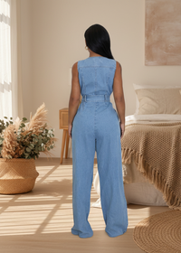 Woman wearing Karina blue denim jumpsuit in a cozy room with a bed and decorative plants.