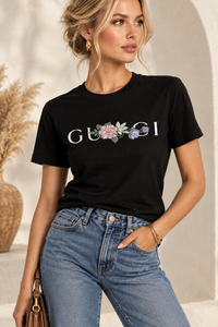 A woman wearing a black short-sleeve t-shirt with a white 'GU*CI' logo and a colorful floral graphic across the chest, paired with blue jeans.