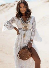 A woman standing on a beach wearing a white Mendez maxi dress with floral patterns, tassel decorations, a V-neckline, and flare sleeves. She is also carrying a brown bag and wearing patterned shoes.