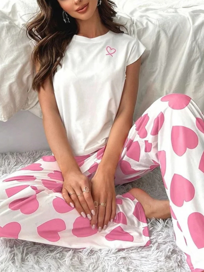 A person wearing a Villa Vibes Passi Princesswhite t-shirt with a heart on it and pink pants with a heart pattern sitting on a bed.