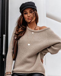 A woman wearing a chunky Peyton knit off-the-shoulder sweater in taupe, a black newsboy cap, and a brown shoulder bag, posing in front of a white wall.