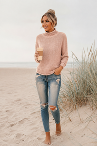Woman in pink Skye sweater and ripped jeans holding a coffee cup on a beach.