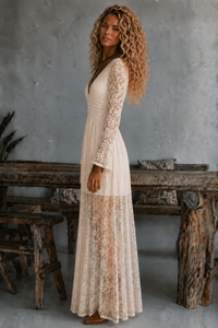 Woman in a long, lace Tessa cream dress standing in a rustic setting with wooden furniture and a gray wall.