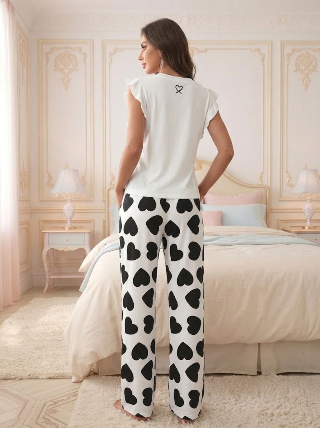 A woman stands in Villa Vibes Passion Princess PJs in bedroom wearing a white sleeveless top and white pants with a black heart pattern, shown from the back.