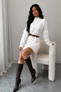 Woman in a Villa Vibes Tanner white sweater dress and brown knee-high boots sitting on a chair.