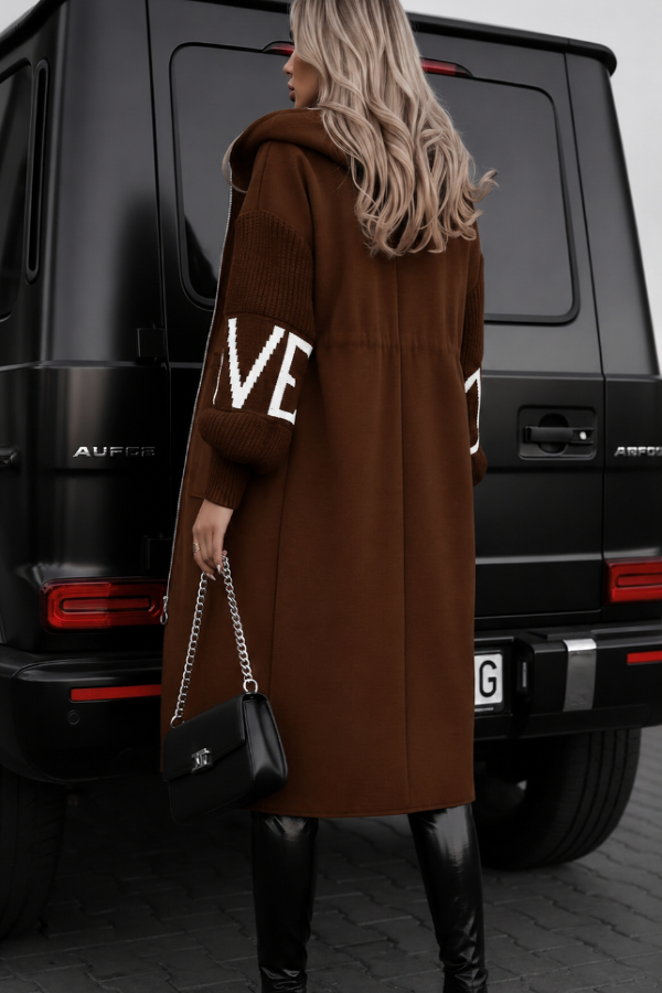 Person wearing a Villa Vibes Kelly  brown coat with 'LOVE' text, standing next to a black luxury SUV.