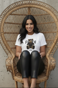 Woman wearing a Villa Vibes designer white t-shirt with a teddy bear graphic and 'Fendi' branding, sitting on a wicker chair.