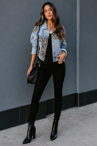 Woman wearing a Villa Vibes Kia denim trucker jacket with leopard print accents, black pants, and black boots against a gray wall.