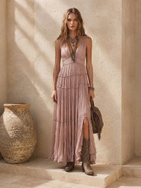 A woman stands on stone steps wearing a Villa Vibes Rya sleeveless, tiered maxi dress in a dusty blush pink color, paired with brown boots and carrying a brown fringed handbag. The setting features textured beige stone walls and a large rustic clay pot.