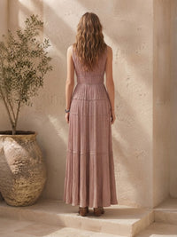 A woman stands with her back to the camera wearing a Villa Vibes Rya sleeveless, tiered maxi dress in a dusty rose pink color, set against a warm beige stone wall with a large potted plant on the left.