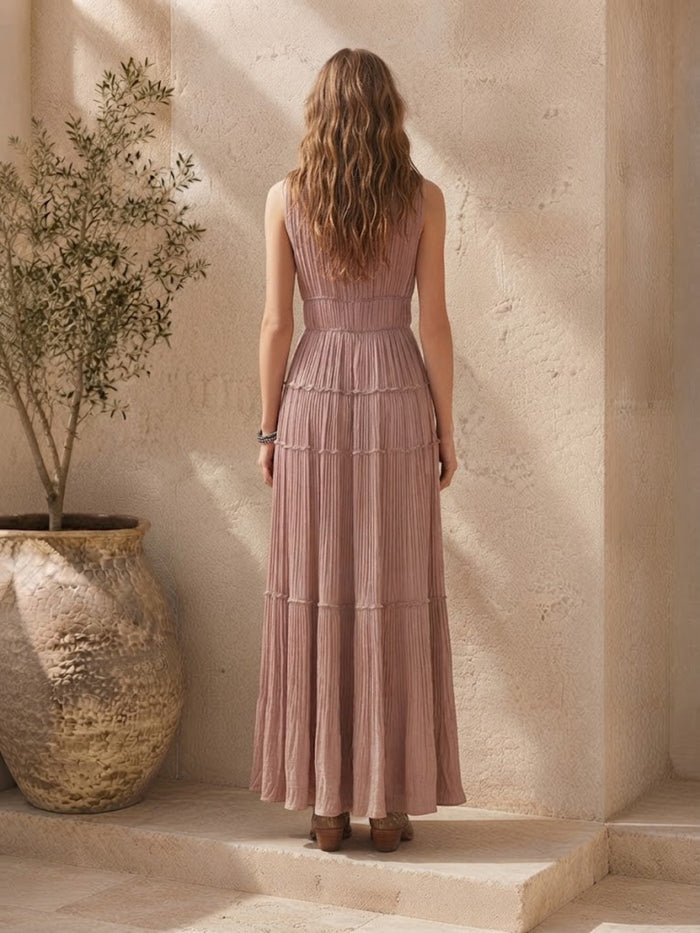 A woman stands with her back to the camera wearing a Villa Vibes Rya sleeveless, tiered maxi dress in a dusty rose pink color, set against a warm beige stone wall with a large potted plant on the left.