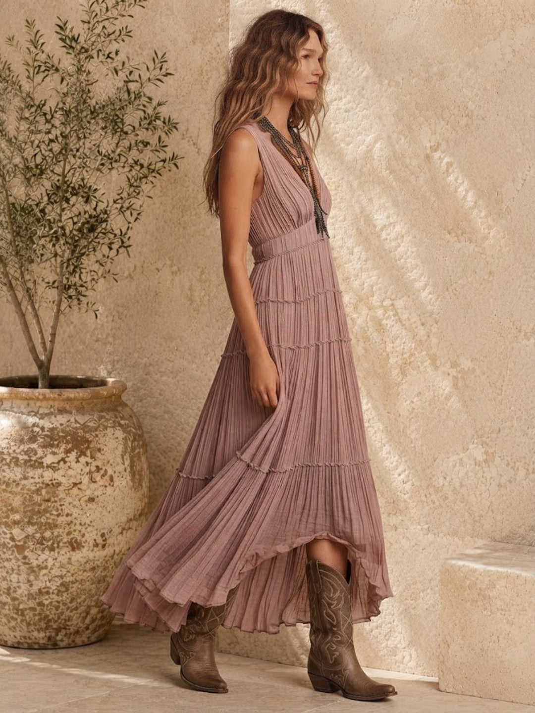 A woman wearing a Villa Vibes Rya sleeveless, tiered midi dress in dusty mauve-pink, paired with brown cowboy boots, standing in a rustic setting with a textured beige wall and a large potted plant.