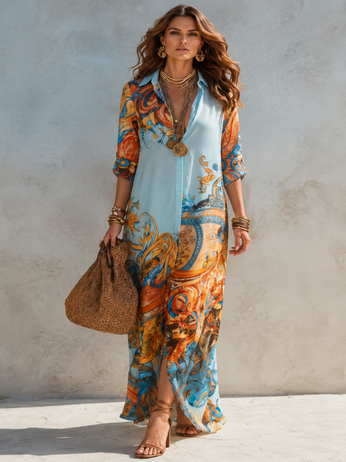 A woman models a Villa VIbes Alanna light blue maxi shirt dress with a vibrant orange, blue, and gold baroque-style print. She is accessorized with gold jewelry, a woven straw bag, and brown strappy sandals, standing against a textured gray concrete wall.