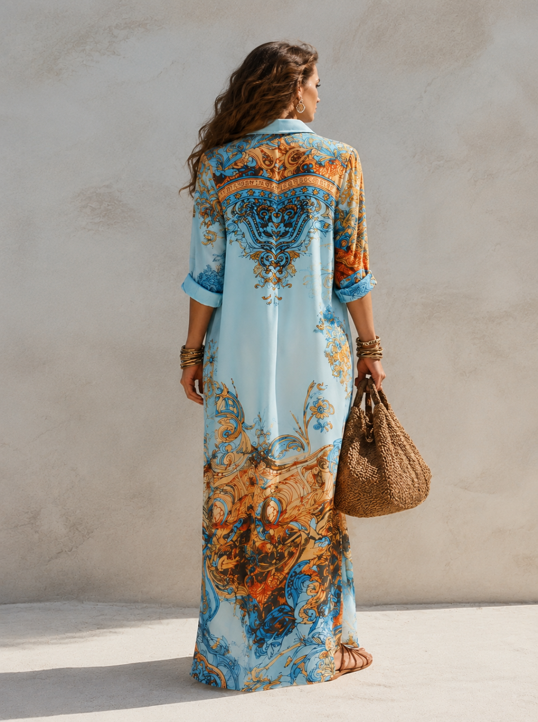 A woman stands with her back to the camera wearing a Villa Vibes Alanna long light blue kaftan dress with an ornate orange and gold baroque-style print. She is holding a brown woven tote bag against a textured beige stucco wall.