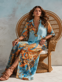 A woman models a Villa Vibes Alanna light blue shirt and matching wide-leg skirt set with an ornate gold and orange baroque print, seated in a large woven rattan peacock chair against a textured light blue wall.
