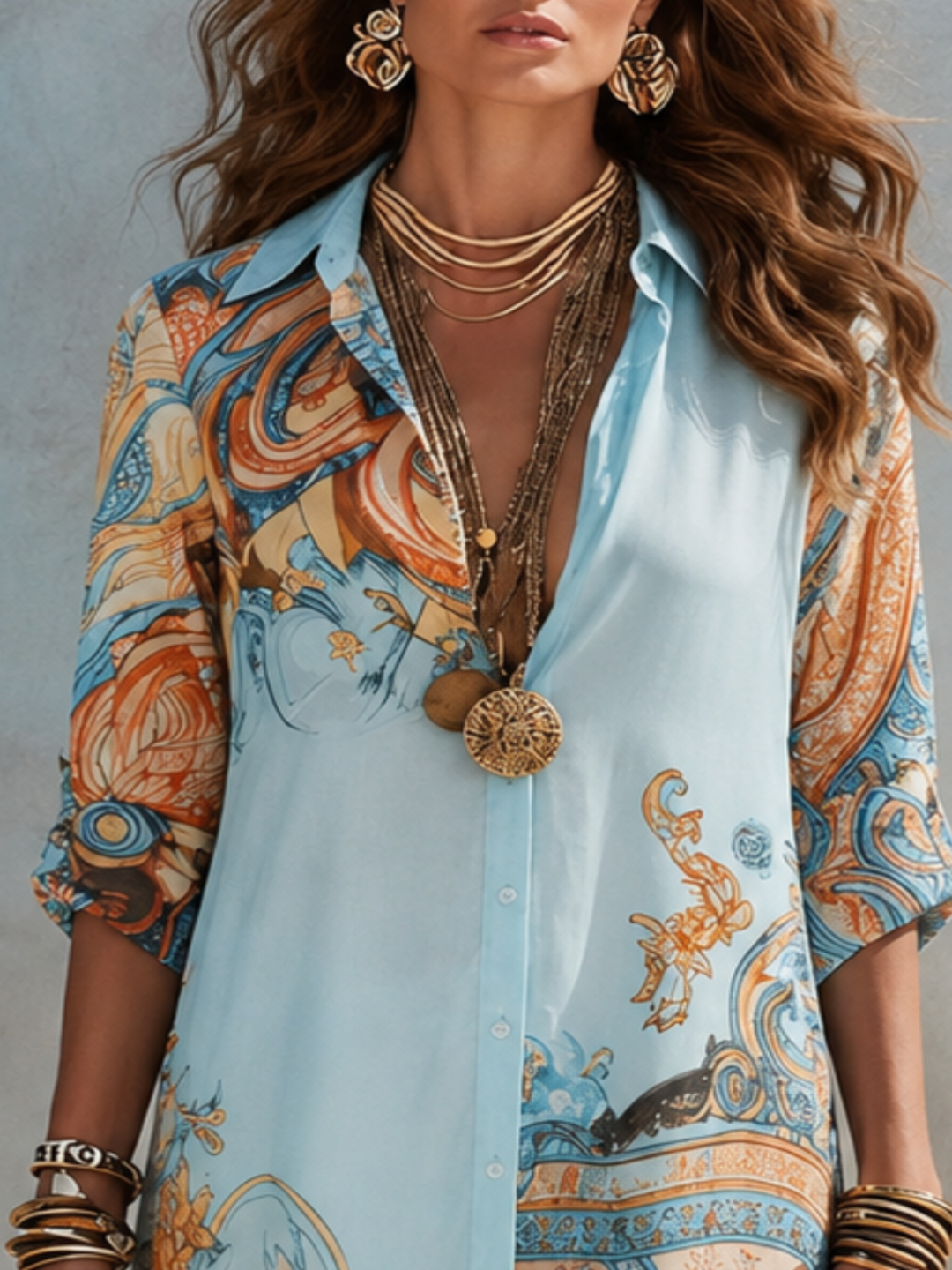 A woman wearing aVilla Vibes Alanna light blue button-up shirt dress with a colorful orange, blue, and beige paisley print. She is accessorized with layered gold necklaces, bracelets, and statement earrings against a textured gray background.