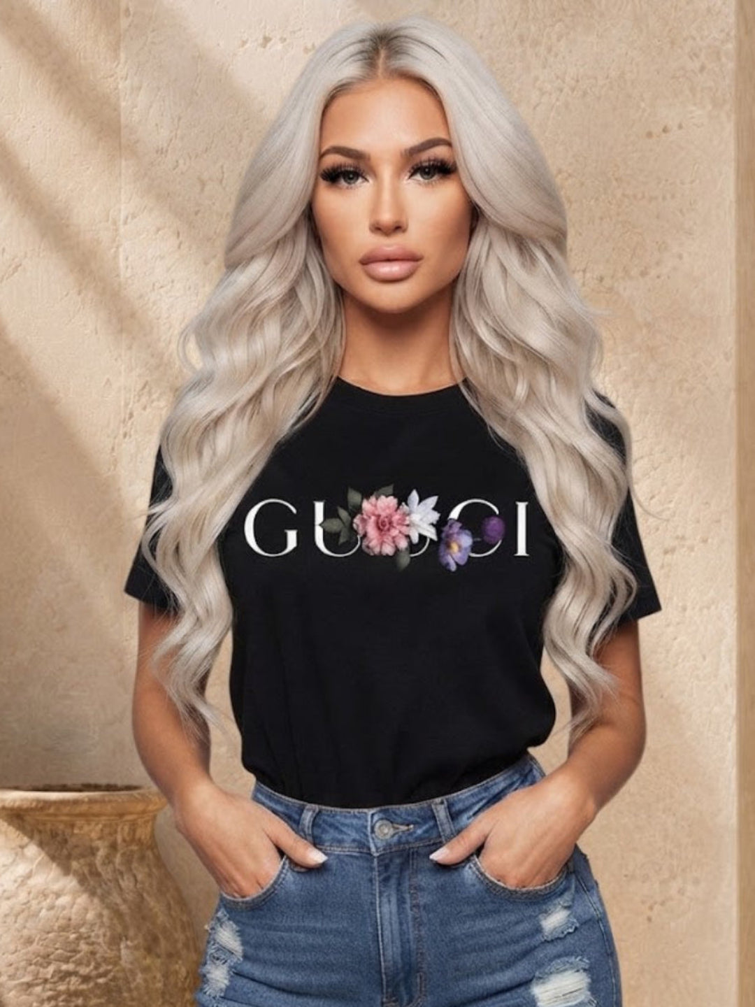 A woman with long platinum blonde wavy hair models a black short-sleeve t-shirt featuring a 'GUCCI' logo printed in white with a pink and purple floral graphic in the center. She is wearing distressed blue jeans and standing in front of a beige stone wall with a decorative ceramic pot.