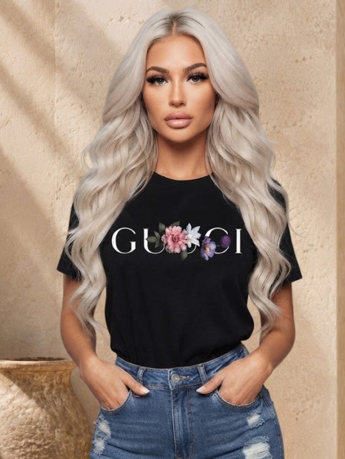 A woman with long platinum blonde wavy hair models a black short-sleeve t-shirt featuring a 'GUCCI' logo printed in white with a pink and purple floral graphic in the center. She is wearing distressed blue jeans and standing in front of a beige stone wall with a decorative ceramic pot.